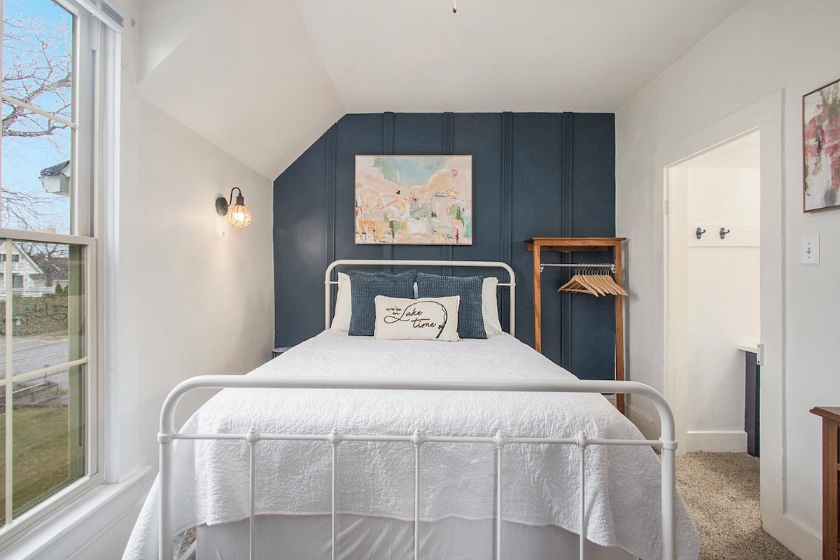 A bedroom with a bed, a window, a wall-mounted light, a painting, and a door.