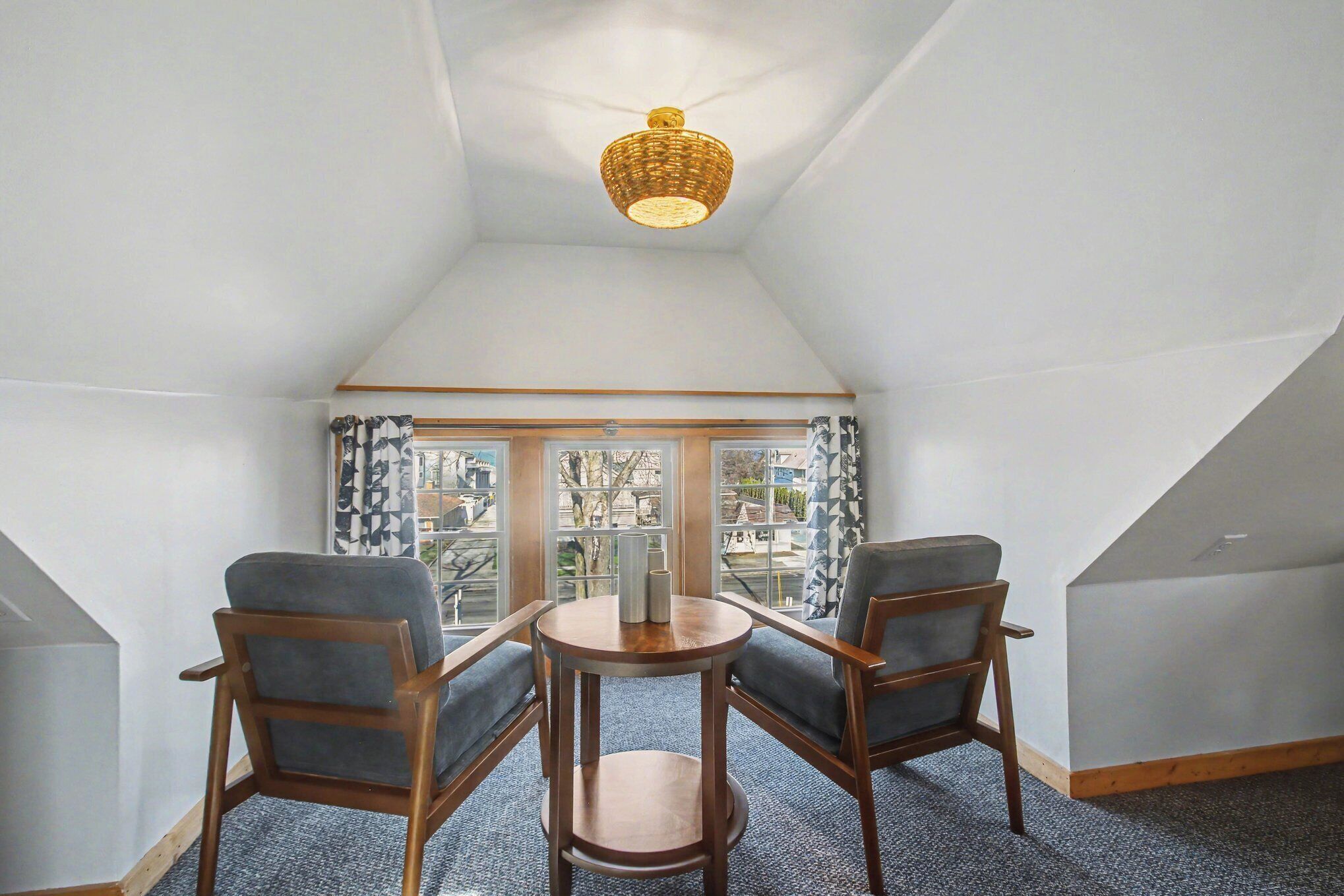 A room with a sloped ceiling, a wicker ceiling light, two wooden chairs with grey cushions, a small wooden table, and a view through a window with patterned curtains.