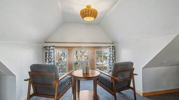 A room with a sloped ceiling, a wicker ceiling light, two wooden chairs with grey cushions, a small wooden table, and a view through a window with patterned curtains.