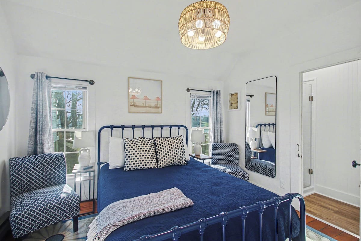 A bedroom with a large bed, a chair, a bedside table, a mirror, and a window with curtains.
