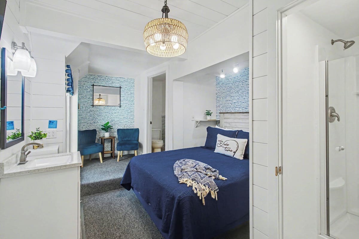 A bedroom with a bed, a desk, a chair, a sink, and a shower.