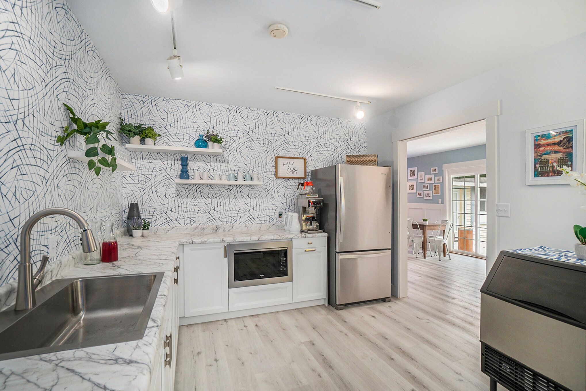 Una cocina moderna con armarios blancos, un refrigerador de acero inoxidable y un microondas. Se observan estantes con plantas y objetos decorativos, además de un lavamanos con grifo.