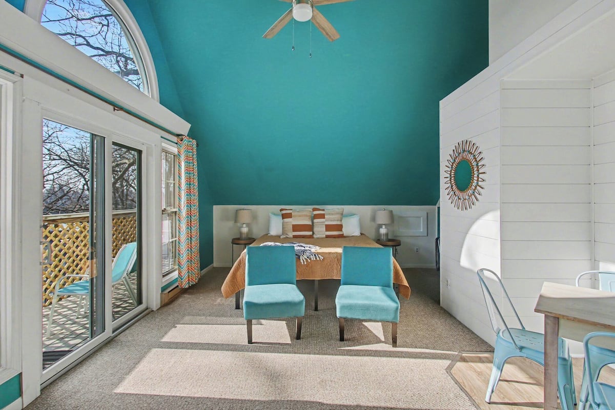 A room with a bed, two chairs, a table, a ceiling fan, and a view through a window.