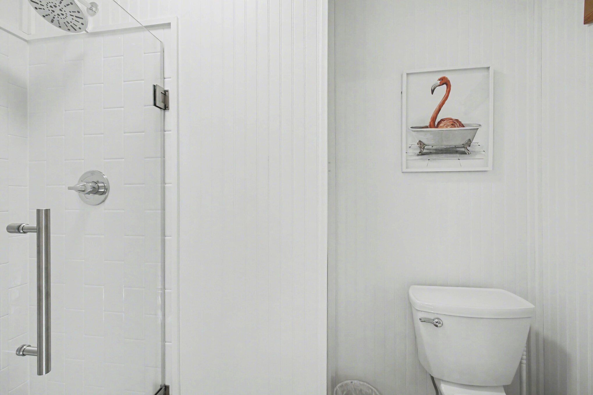 A bathroom with a white tiled wall, a white toilet, a shower with a glass door, and a framed picture of a flamingo in a bathtub.