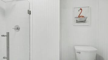 A bathroom with a white tiled wall, a white toilet, a shower with a glass door, and a framed picture of a flamingo in a bathtub.