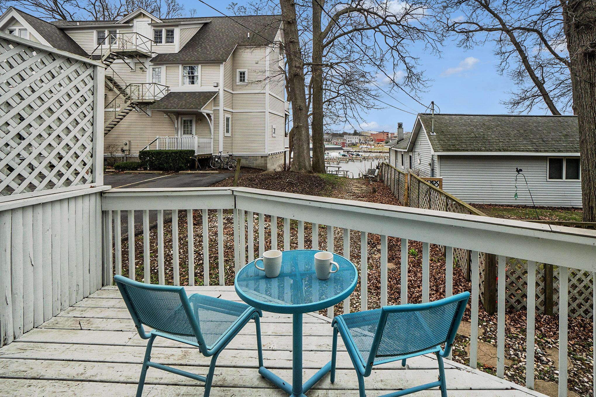 Une terrasse en bois avec deux chaises bleues et une petite table, donnant sur un quartier résidentiel avec des maisons et des arbres.