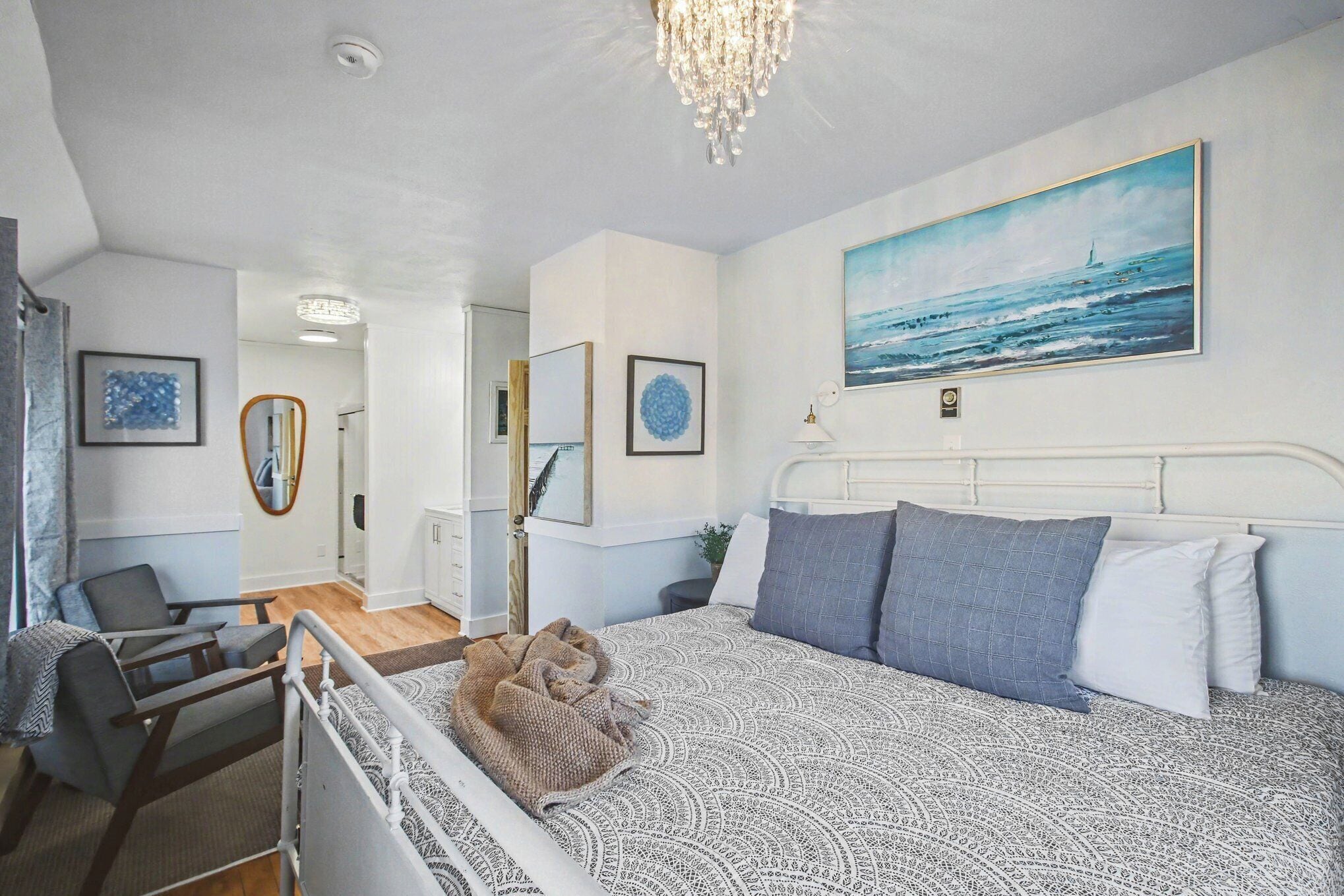 A bedroom with a bed, a chair, a mirror, and a painting of the sea.