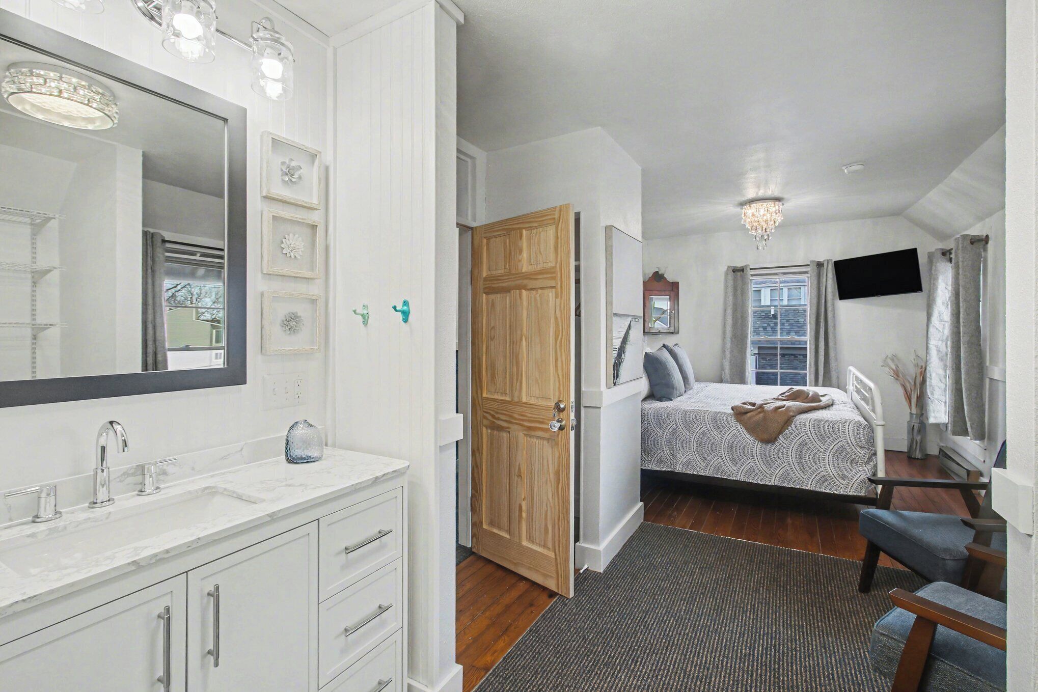 A bedroom with a bed, a TV, a wooden door, and a bathroom with a large mirror and sink.