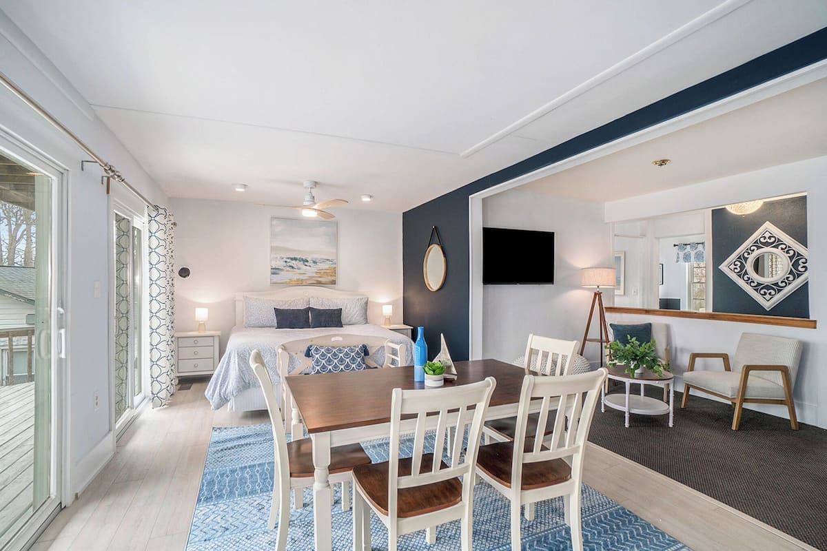 A modern bedroom with a large bed, a dining table with chairs, a small side table, and a mirror on the wall.