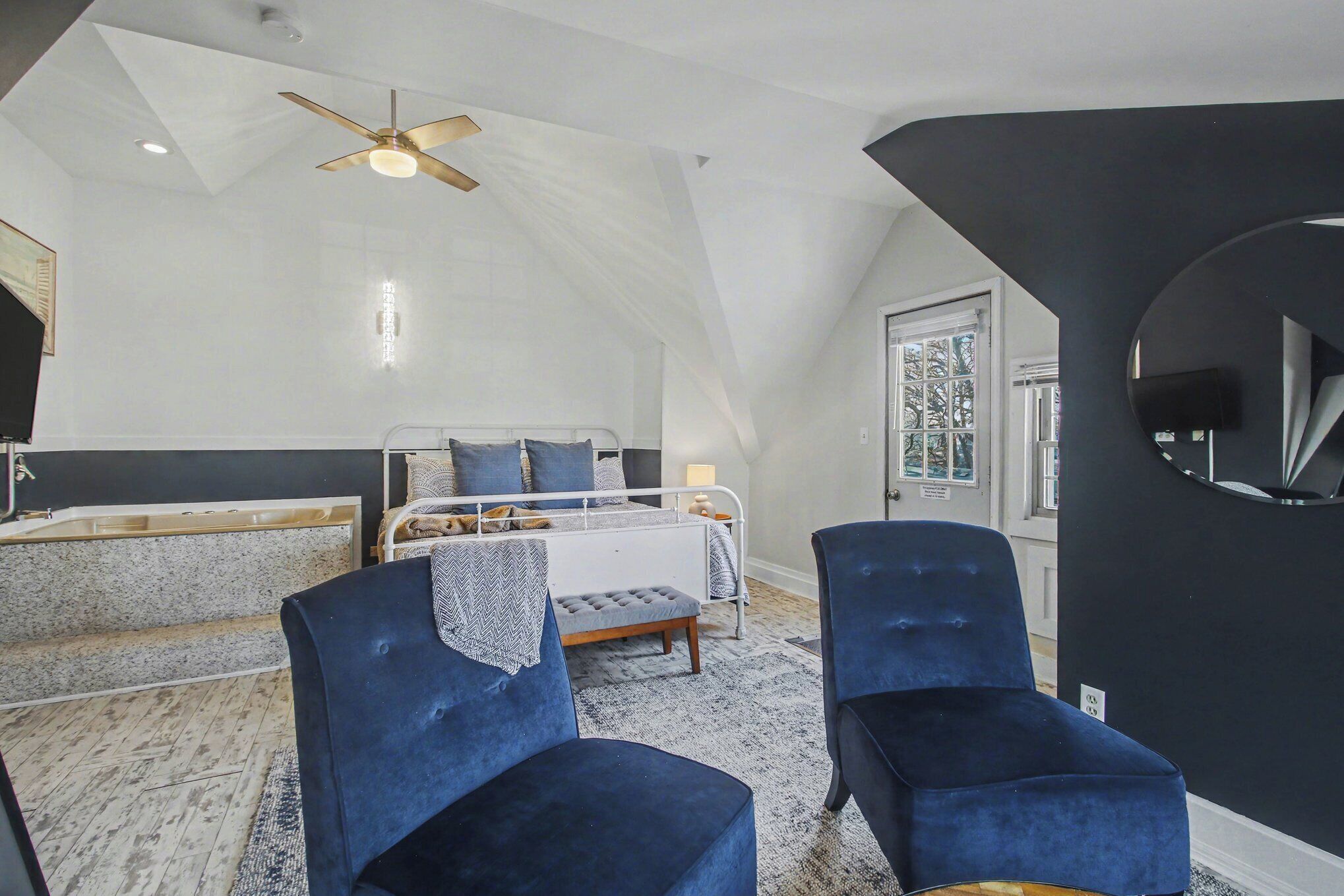 A bedroom with a sloped ceiling, a bed, two blue chairs, a ceiling fan, and a window.