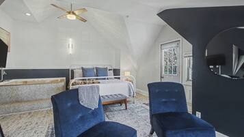 A bedroom with a sloped ceiling, a bed, two blue chairs, a ceiling fan, and a window.