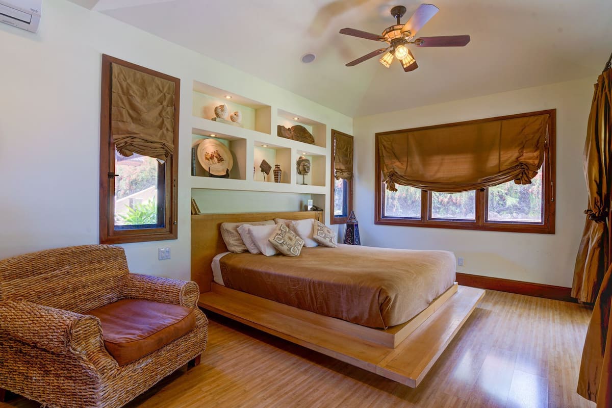 A bedroom with a bed, a wicker chair, a ceiling fan, and shelves with decorative items.