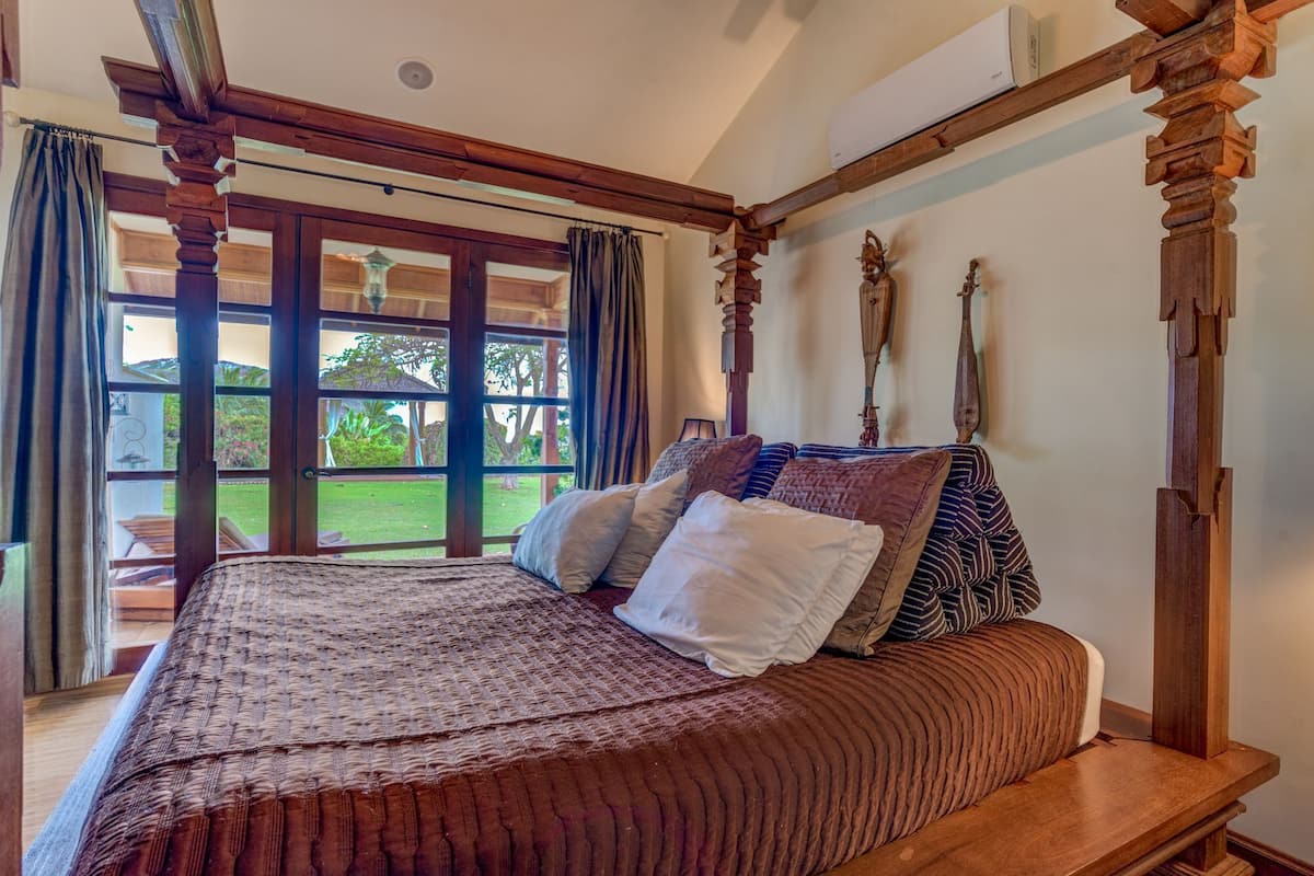 A four-poster bed with a wooden headboard, a large window with curtains, and a view of a garden.