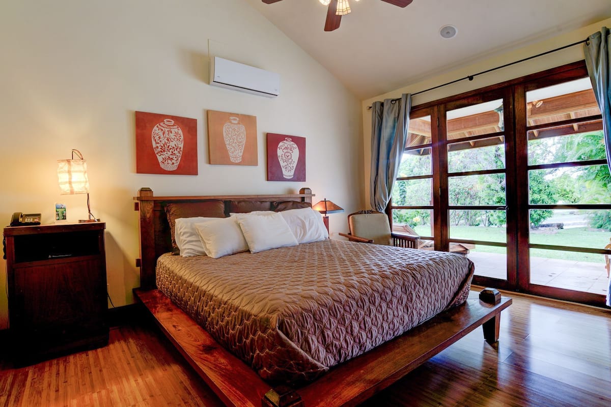 A bedroom with a wooden bed, a bedside table with a lamp, and a window with a view of greenery.