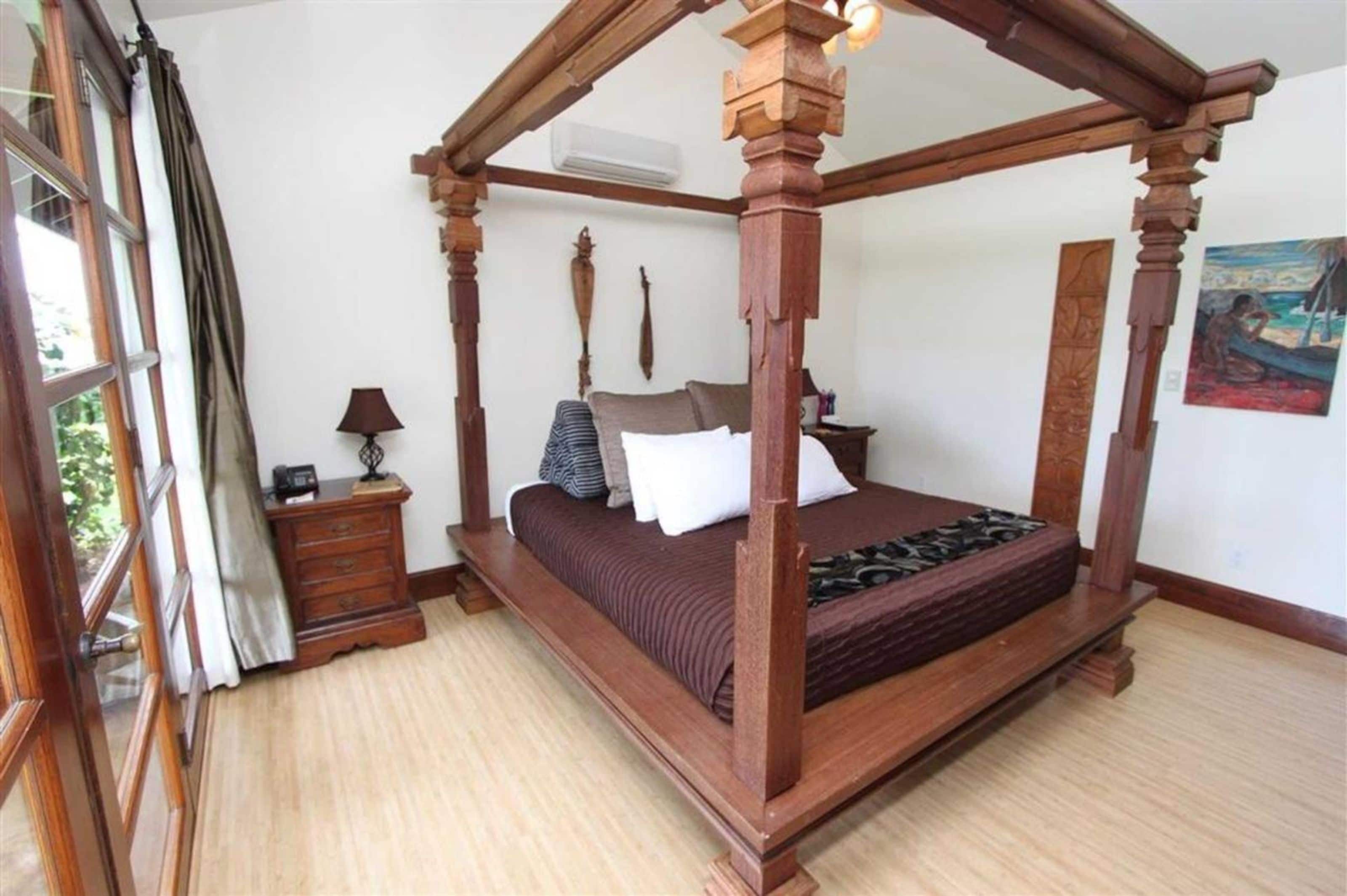 Uma cama de dossel com estrutura de madeira, uma mesa de cabeceira com abajur e um quadro na parede.