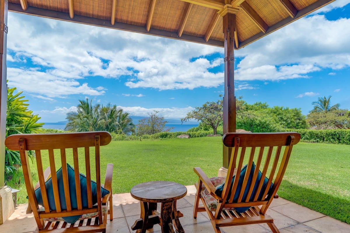 Two wooden chairs with cushions, a small round table, and a covered patio area overlooking a grassy landscape with trees and a view of the ocean.