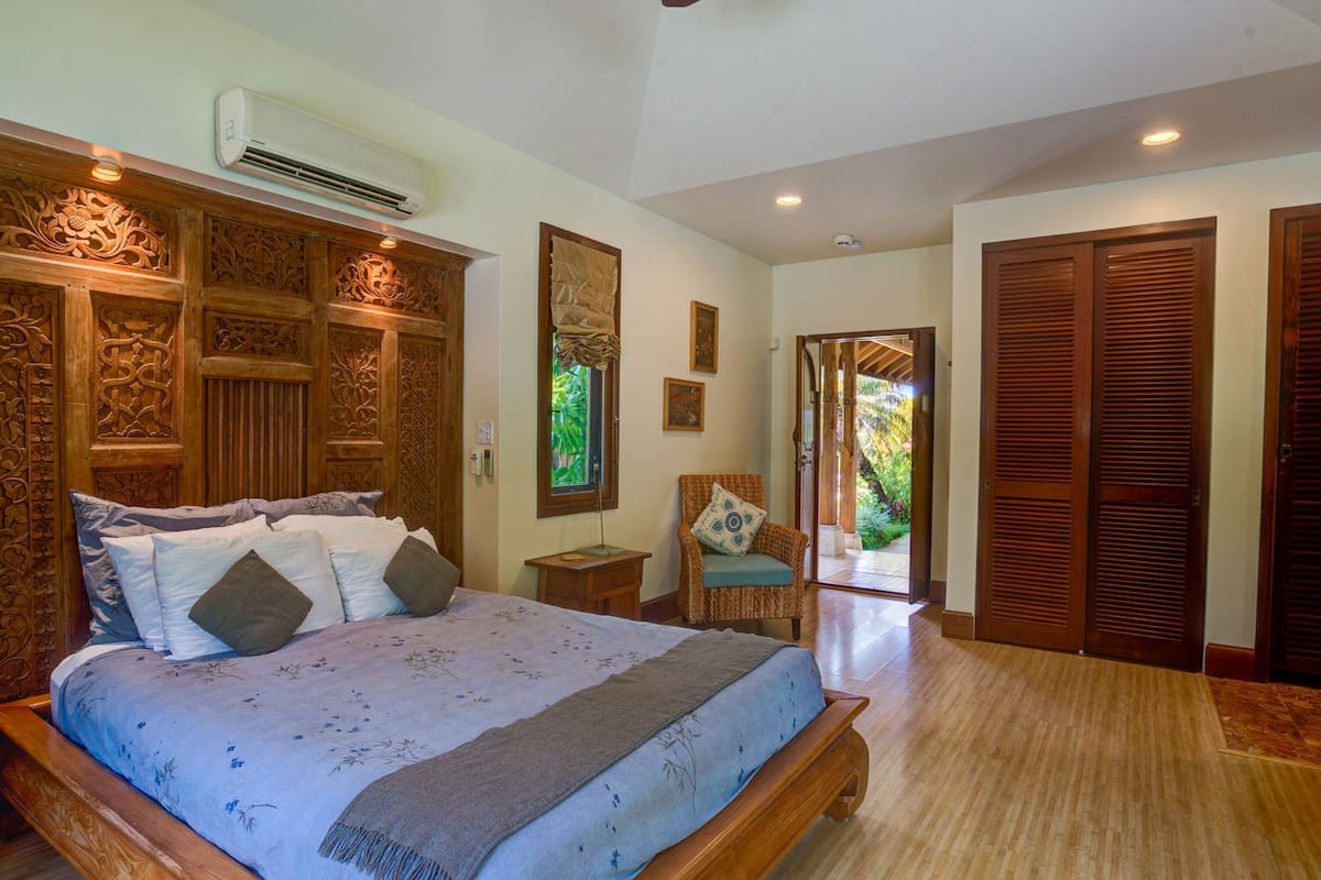 A spacious bedroom with a large bed, wooden paneling, an air conditioning unit, and a view to a garden through a sliding door.