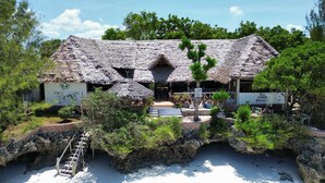 A traditional thatched-roof building with a pool, surrounded by lush greenery and a rocky landscape. - Mbuyuni Beach Village - Bungalows (Jambiani)