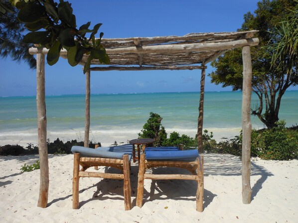 A wooden beach chair and umbrella under a pergola with a view of the ocean.