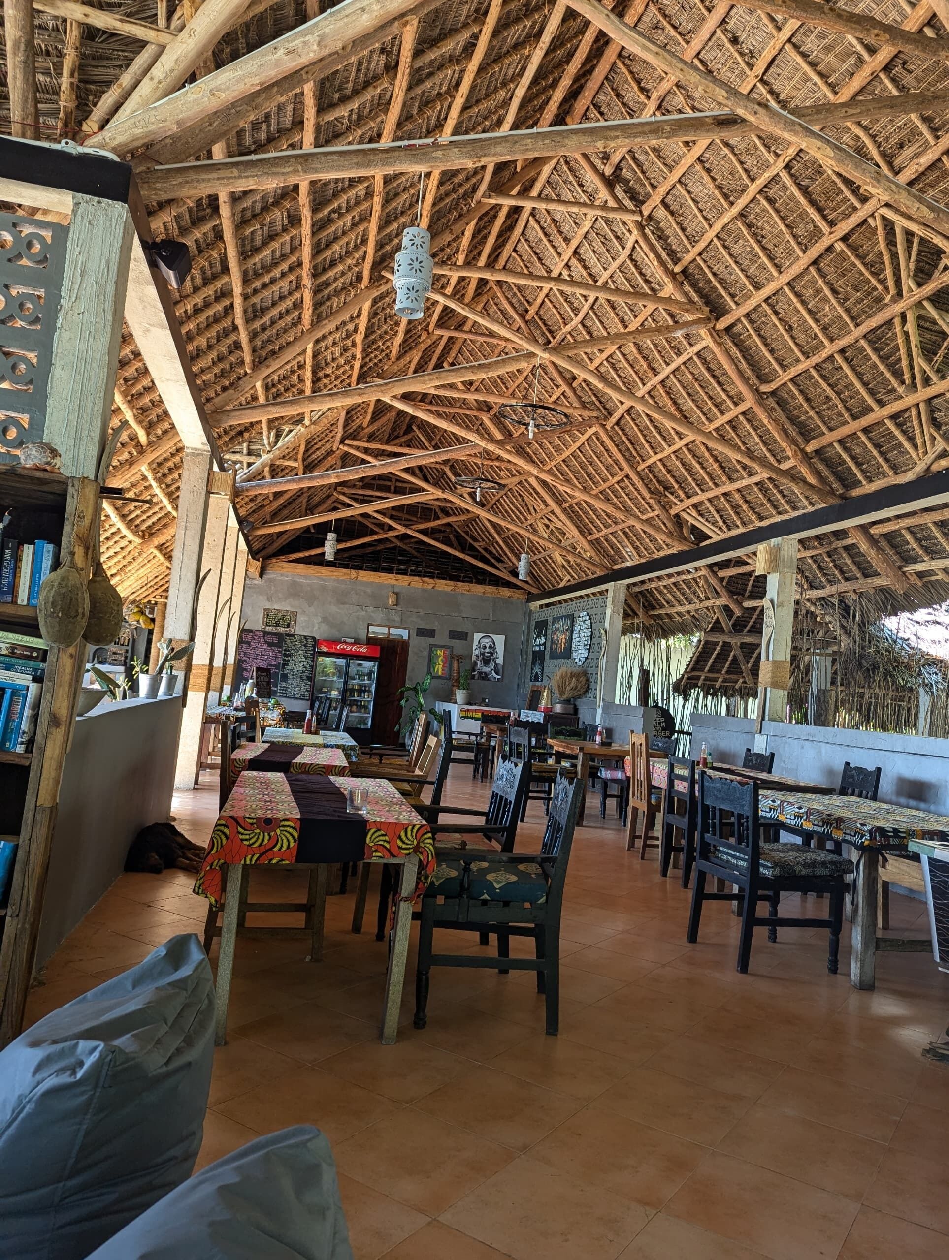 Un comedor interior con un techo alto de paja, mesas y sillas de madera, y suelo de baldosas.