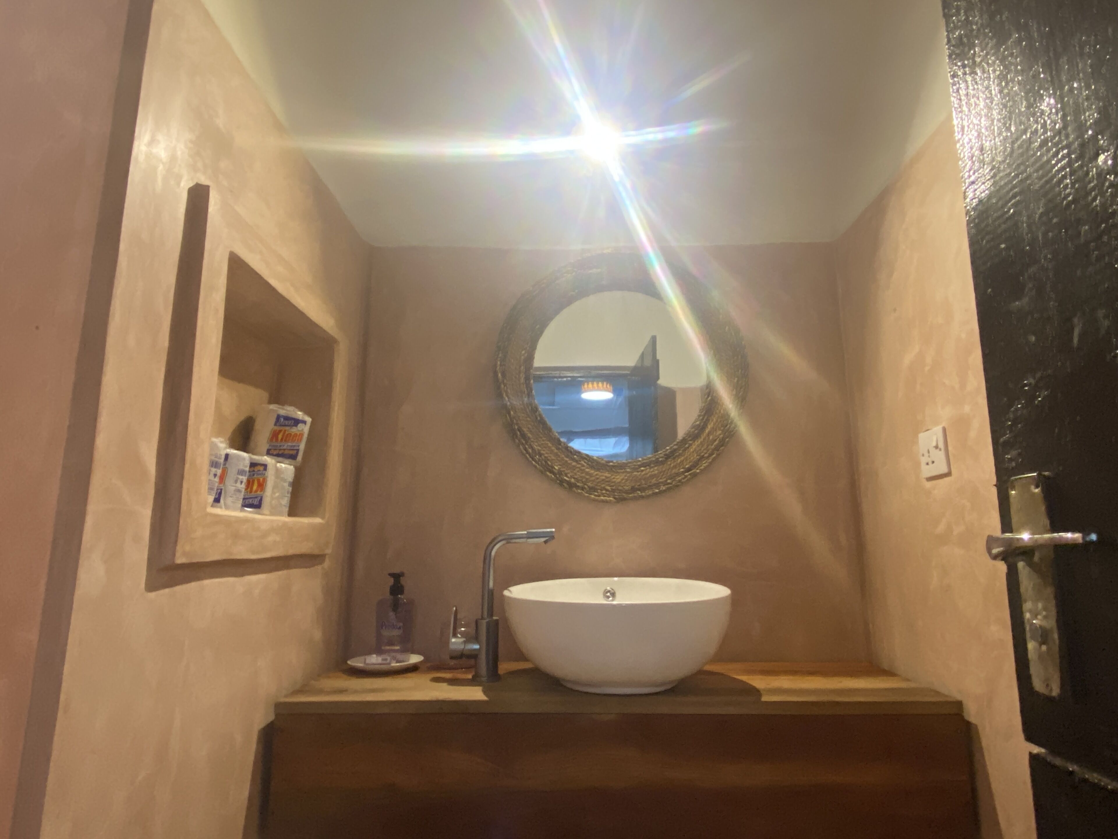 Ein Badezimmer mit einem weißen Waschbecken, einem runden Spiegel, einer Ablage mit Toilettenartikeln und einer Tür mit schwarzem Rahmen.