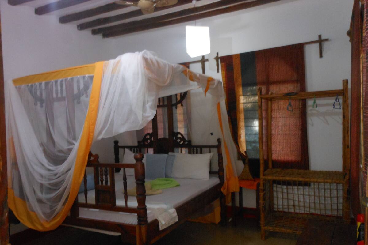 Una cama individual con dosel, una silla de madera, una mesita y una ventana con cortinas.