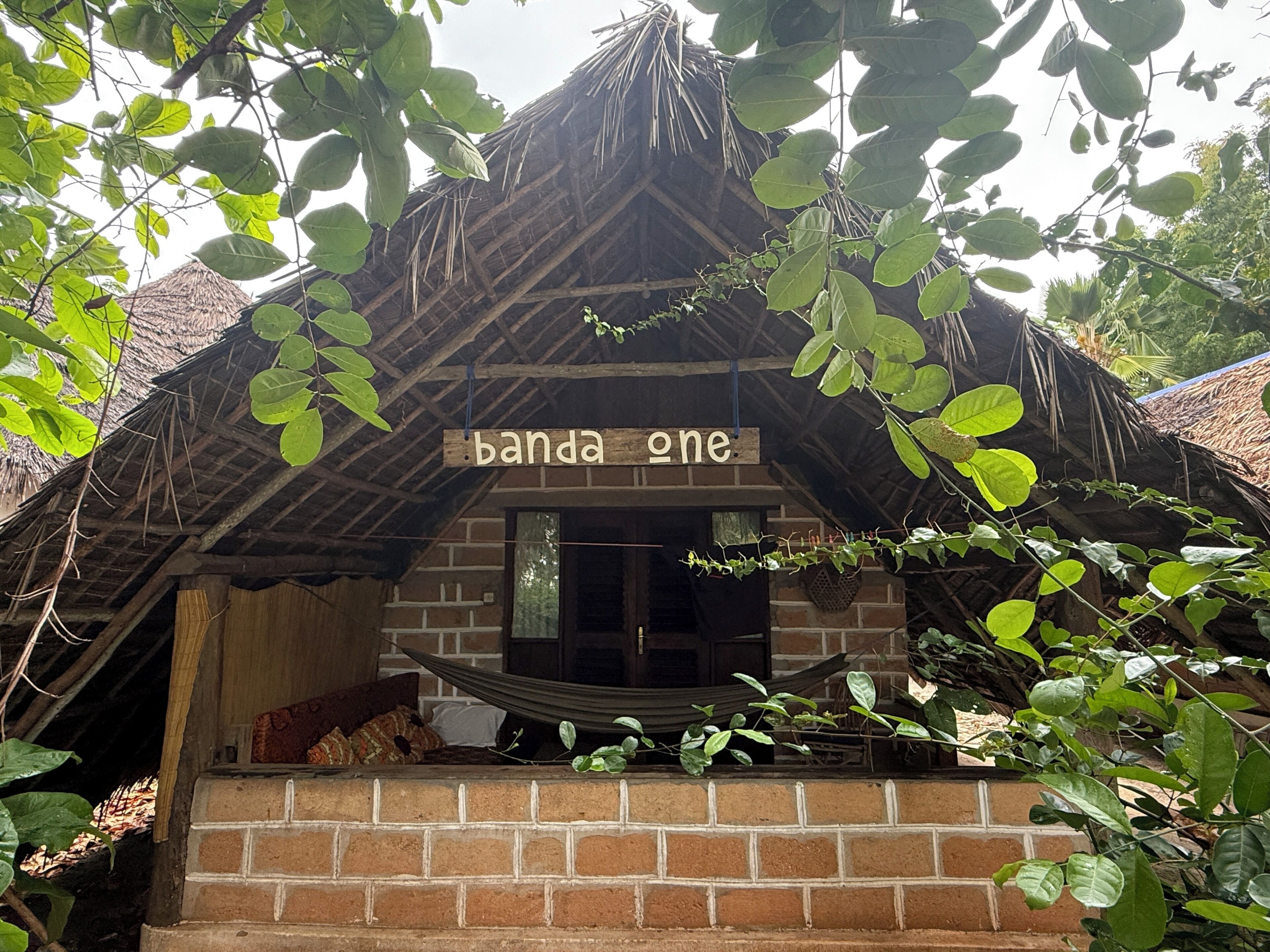 A rustic hut with a thatched roof, a wooden sign reading 'banda one,' and a brick wall.