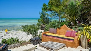 A beachside lounge with a sofa and a small table, surrounded by tropical plants and overlooking the ocean. - Mbuyuni Beach Village - Bungalows (Jambiani)