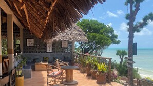 An outdoor seating area with a thatched roof, wooden chairs, and potted plants. - Mbuyuni Beach Village - Bungalows (Jambiani)