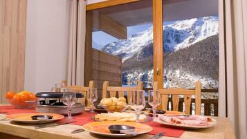 A dining table set with plates, glasses, and a basket of oranges, with a mountain view through a window.