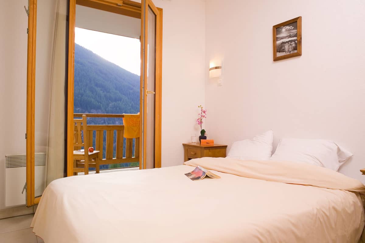 Une chambre à coucher avec un lit, une table de chevet, un tableau encadré et une vue sur une montagne, visible à travers une porte ouverte.