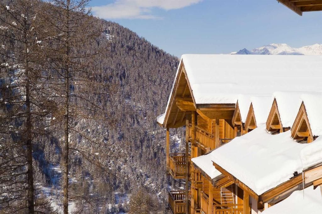 Una cabaña de madera con techo nevado, con montañas al fondo.