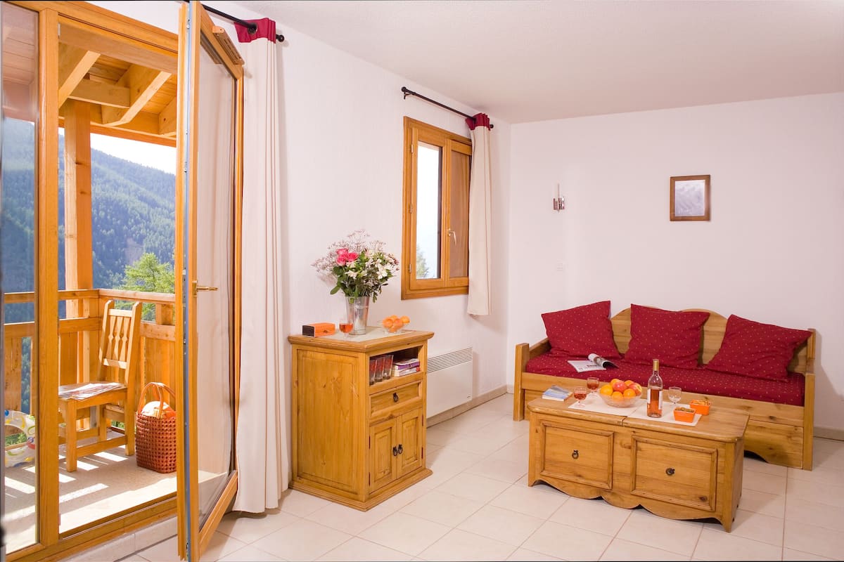 Une pièce avec un canapé en bois, un meuble en bois, une table à manger ornée d’un vase de fleurs et une vue sur une montagne, visible à travers une porte coulissante.