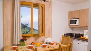 A wooden dining table with breakfast items, a view of mountains through a window, and a kitchen with a microwave. - Madame Vacances - Residence Les Balcons des Airelles (Les Orres)