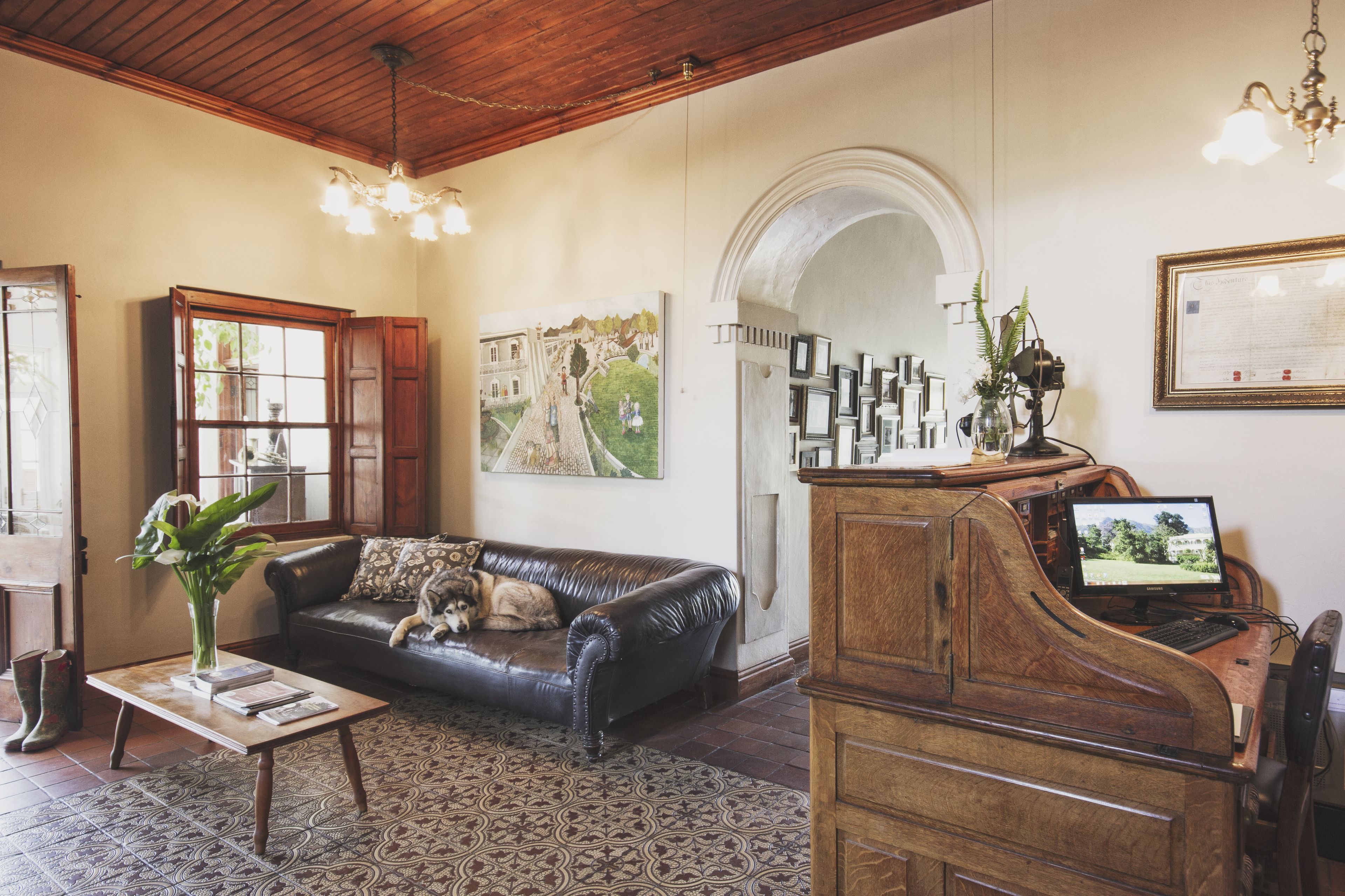 A living room with a black leather sofa, a wooden coffee table, a painting on the wall, a piano, and a chandelier.