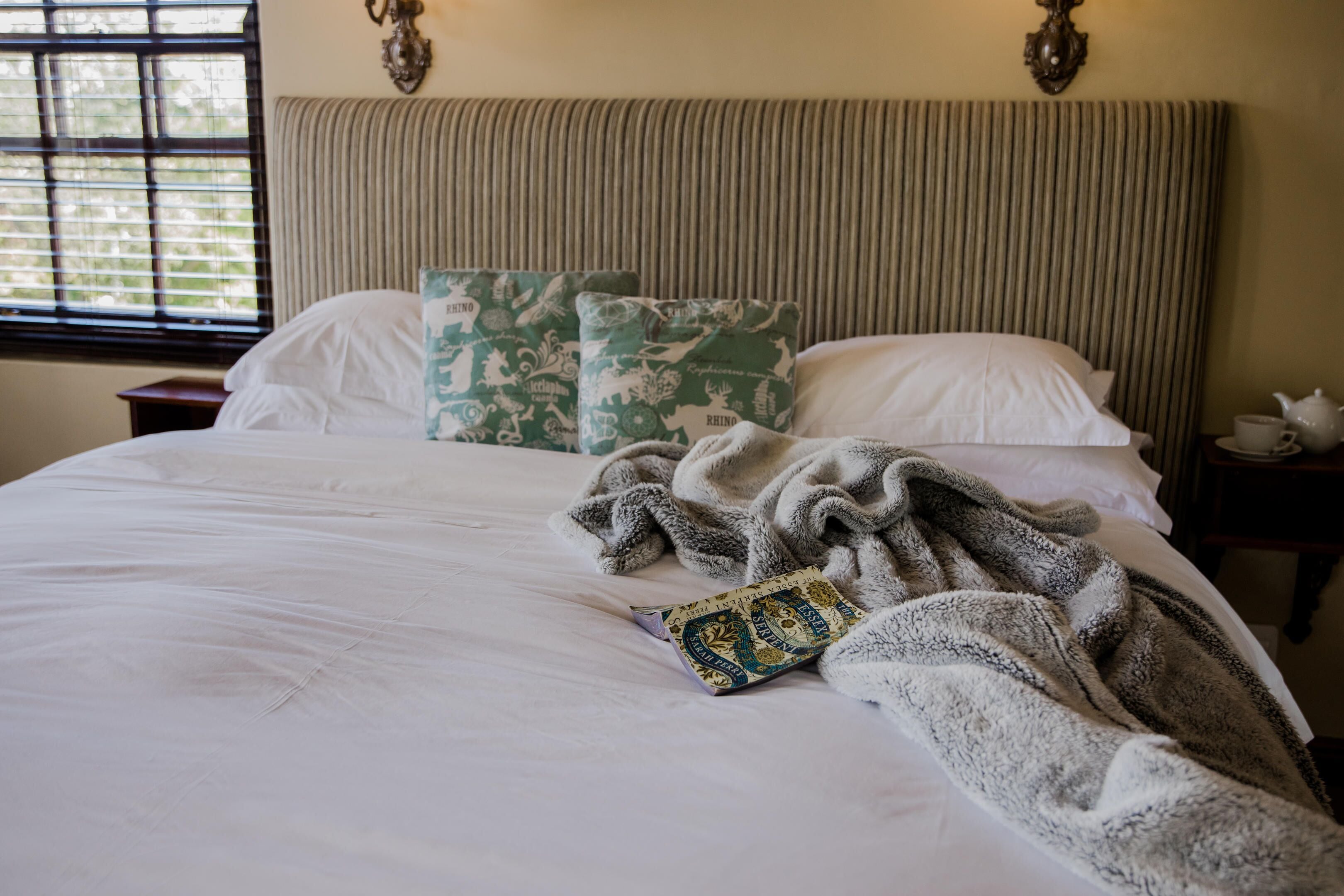 Een net opgemaakt bed met een boek en een plaid, een gestreept hoofdeinde en een raam met blinds.