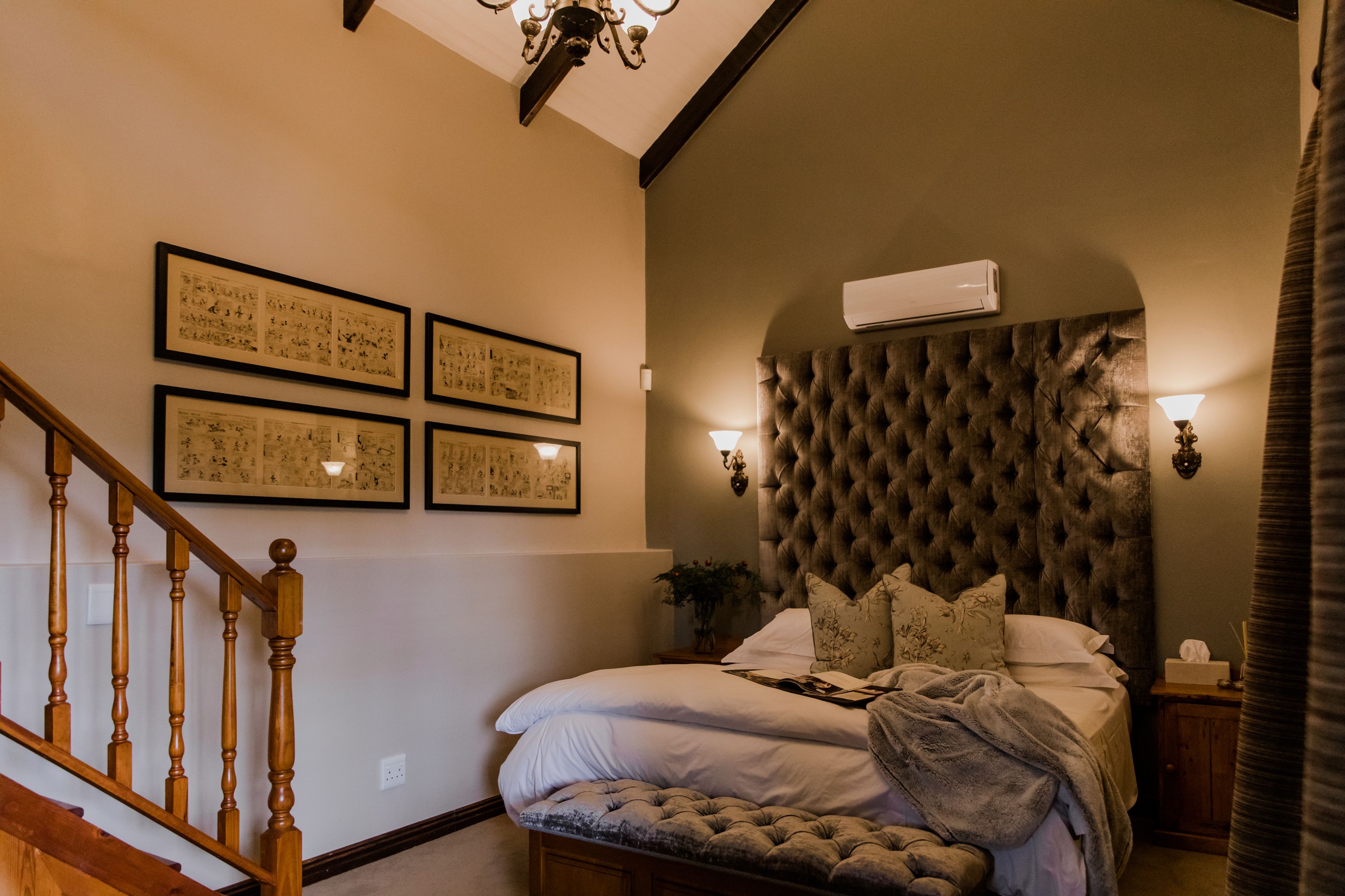 A bedroom with a tufted headboard, a wooden staircase, and framed artwork on the wall.