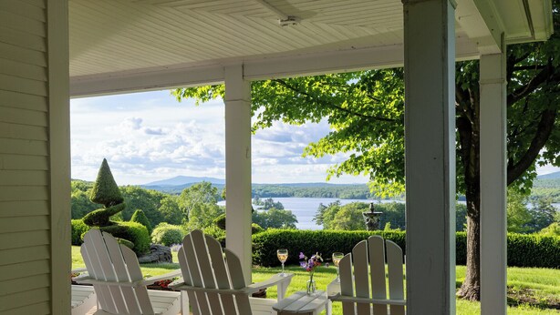 Une véranda abritée, dotée de fauteuils Adirondack et d’une table avec des verres à vin, offrant une vue sur un lac et une végétation luxuriante.