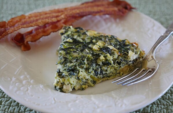 Una porción de quiche de espinacas y queso sobre un plato blanco, con un tenedor, acompañada de tiras de tocino.