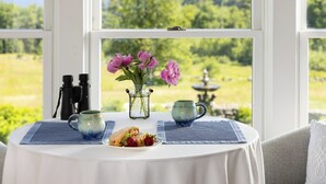 Une table sur laquelle se trouvent une assiette de fraises, une tasse et des jumelles, le tout placé devant une fenêtre donnant sur un paysage verdoyant et un plan d’eau.
