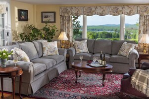 A cozy living room with two grey sofas, a red patterned rug, and a view of a lake and forest through large windows.