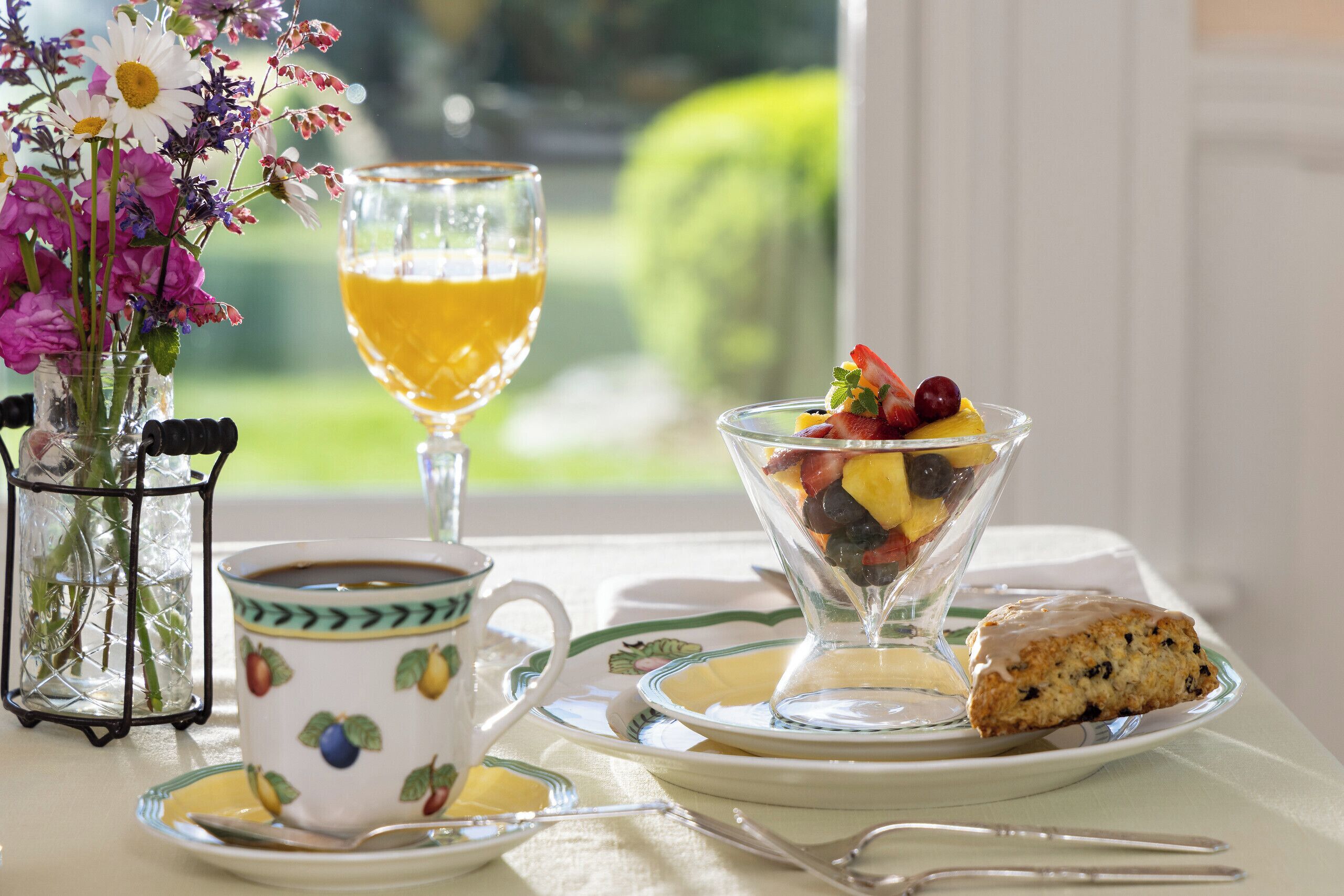 A breakfast setting with a glass of orange juice, a cup of coffee, a bowl of fruit salad, a slice of cake, and a vase of flowers.