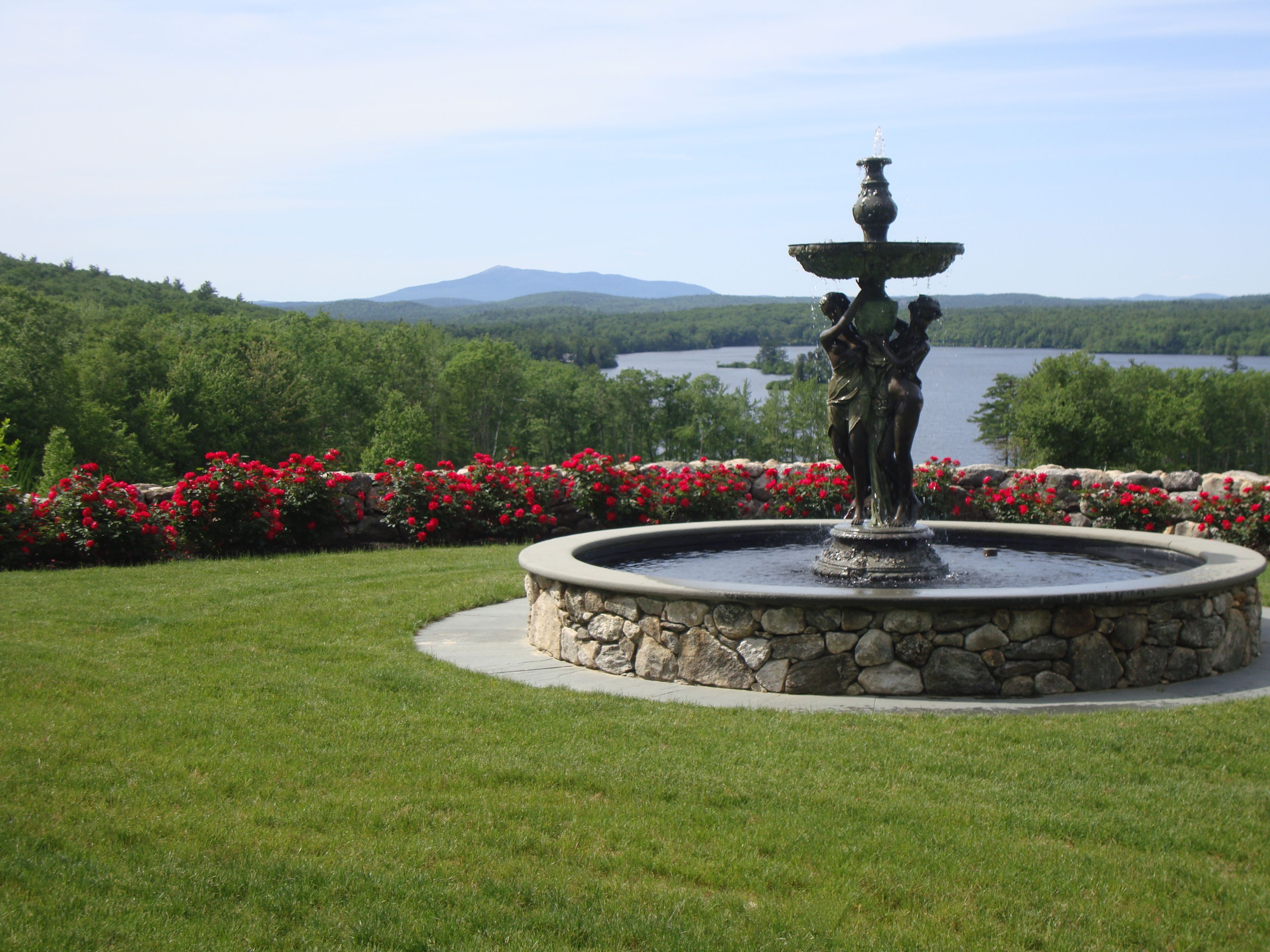 A fountain with a statue in the center, surrounded by a stone circular base and a flower bed with red flowers.
