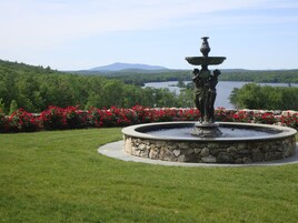 A fountain with a statue in the center, surrounded by a stone circular base and a flower bed with red flowers.