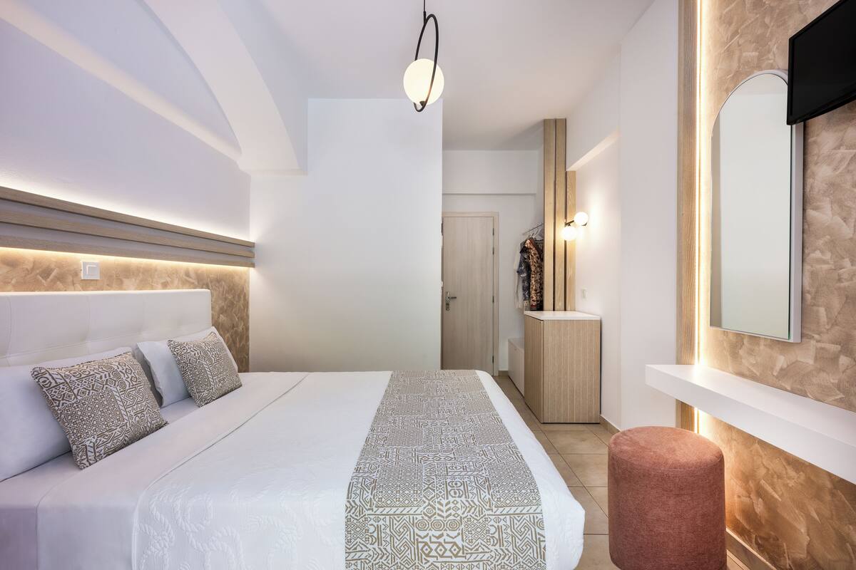 A modern hotel room with a large bed, a bedside table, a mirror, and a small stool.