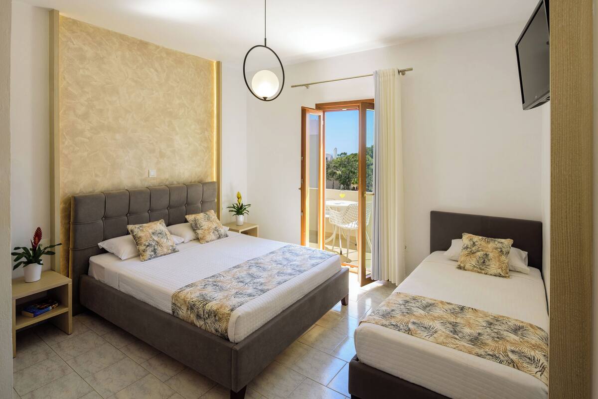 A hotel room with a large bed, a smaller bed, a TV, a bedside table, a potted plant, and a view of the outdoors.