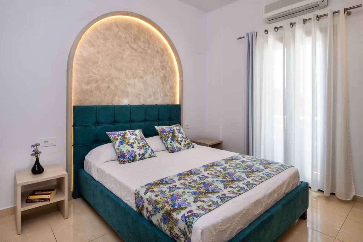 A bedroom with a bed, a nightstand, a vase with flowers, and a wall-mounted air conditioner.
