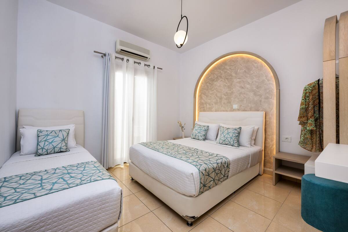 A modern hotel room with two beds, a large arched headboard, a small table, and a blue chair.