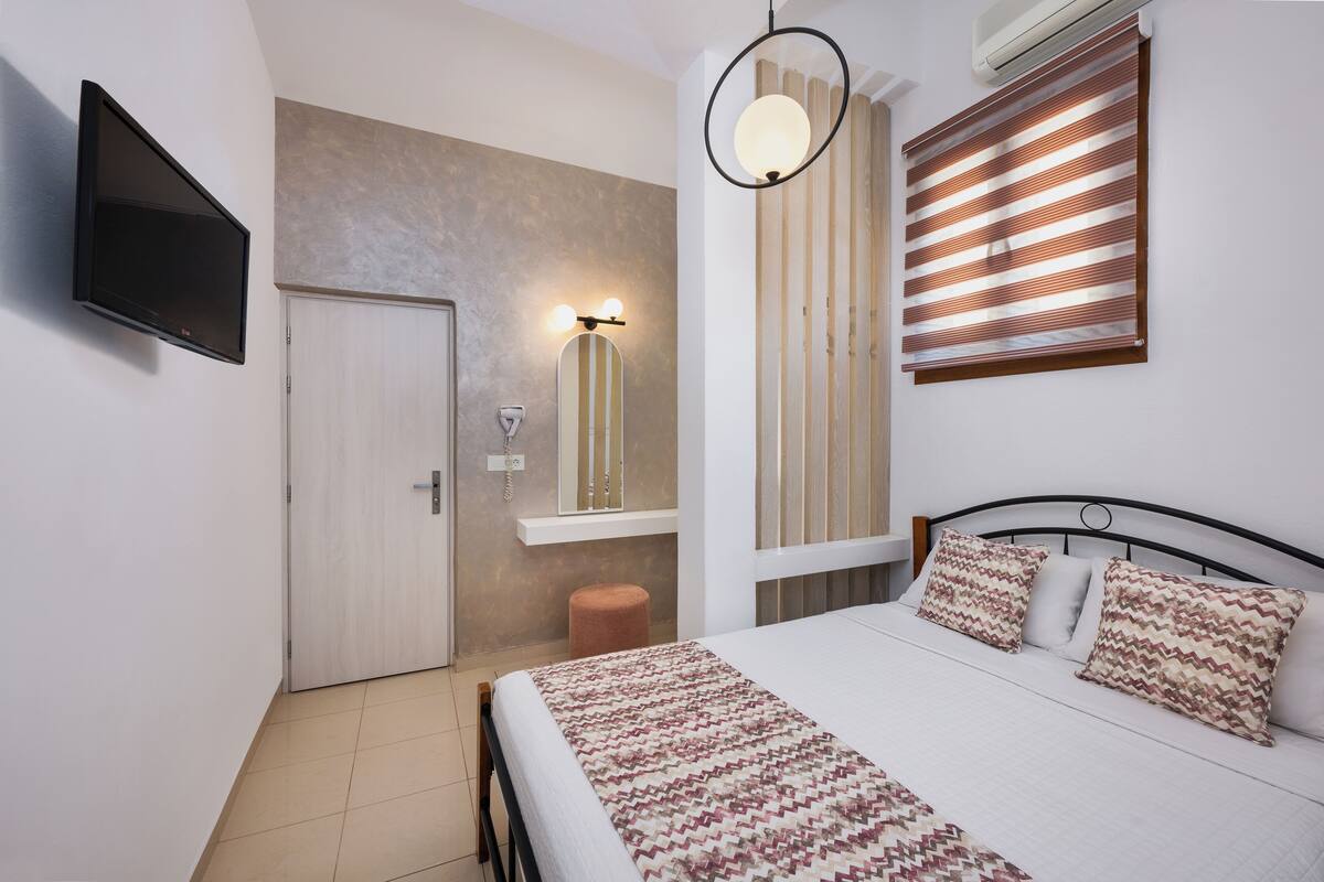 A bedroom with a bed, a TV mounted on the wall, a bedside table, and a window with blinds.