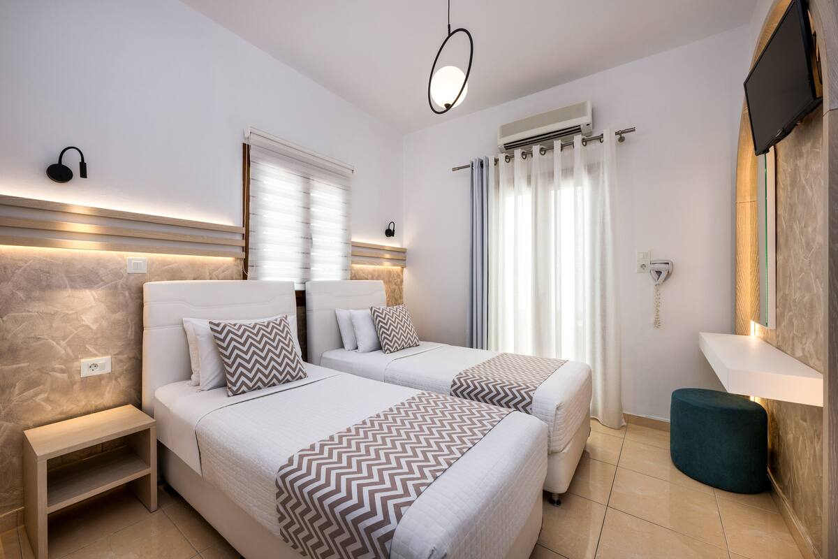 A modern hotel room with two beds, a bedside table, a small stool, a TV mounted on the wall, and a window with curtains.